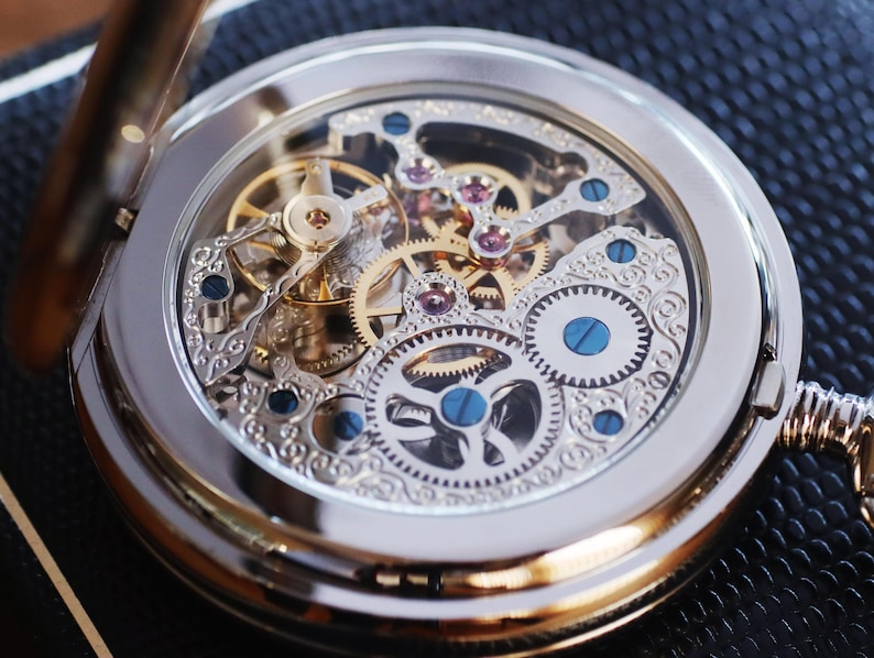 May include: A silver pocket watch with a transparent face, showcasing the intricate gold and silver gears and jewels of the watch mechanism. The watch has a silver chain and a hinged cover. The watch is a classic timepiece.