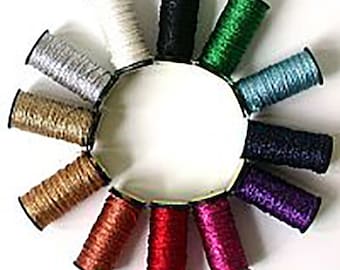 May include: A collection of spools of decorative cord in various colors, including silver, gold, black, green, and red. The spools are arranged in a circular pattern around a white center, showcasing the variety of colors available.