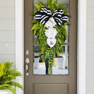 May include: A black and white striped bow with a white heart sign that says "Home Est. 2023" hangs on a brown door with a green leafy garland.