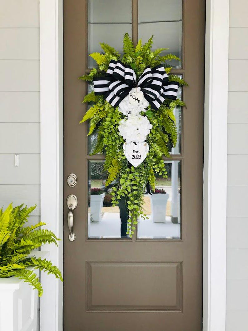 May include: A black and white striped bow with a white heart sign that says "Home Est. 2023" hangs on a brown door with a green leafy garland.