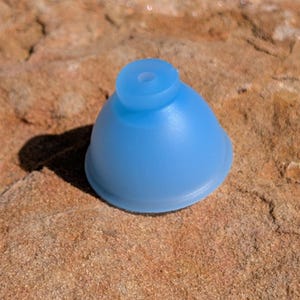 May include: A translucent light blue silicone suction cup with a smooth, dome-shaped body and a small circular opening at the top. The object features a wide, flat base rim designed for creating a vacuum seal on smooth surfaces.