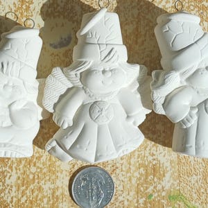 May include: Three unpainted ceramic angel ornaments. Each angel has wings, a hat, and a dress. The ornaments are white and have a loop for hanging. A coin is placed below the ornaments for size comparison.