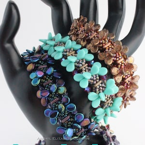 May include: Three beaded bracelets in shades of blue, turquoise, and bronze are displayed on a black hand. The bracelets feature a floral design with iridescent and metallic beads. The text "Butterfly Fields Forever Bracelet Tutorial" is visible.