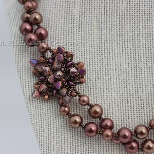 May include: A handcrafted necklace featuring a cluster of iridescent beads and a double strand of brown-purple pearls. The necklace has a central focal point with a starburst design and is displayed on a neutral background.