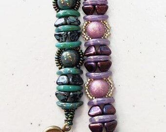 May include: Two beaded bracelets. One is teal and dark green with a gold clasp. The other is purple and lavender with a silver clasp. The bracelets feature round and oval beads with decorative accents.