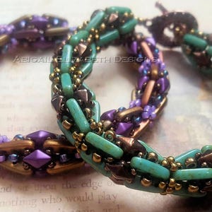 May include: Two beaded bracelets. One is purple, gold, and bronze. The other is turquoise, gold, and bronze. The bracelets are made of small, decorative beads. The text "ABIGAIL ELIZABETH DESIGNS" is visible.