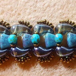 Op de afbeelding: Een kralenarmband met een herhalend patroon van iriserende glazen en turquoise kralen. De armband heeft bronzen metalen accenten en wordt gepresenteerd op een neutrale achtergrond.