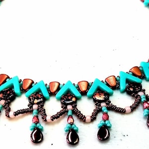 Op de afbeelding: Een kralenketting met turquoise, koperkleurige en donkerbruine elementen. De ketting heeft een herhalend patroon van turquoise pijlen en koperkleurige kralen. Hangende druppelvormige kralen in donkerbruin en kleinere kralen in roze en turquoise maken het ontwerp af.