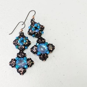 May include: Handmade earrings featuring a cross-shaped design with a large, light blue crystal center. The earrings have a black and copper-colored beaded frame, with smaller blue and green crystals. The ear wires are dark metal.