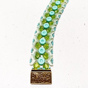 Op de afbeelding: Een kralenarmband met een herhalend patroon van lichtblauwe, groene en witte kralen. De armband heeft een decoratieve bronskleurige sluiting. Het ontwerp bevat kleine, ronde kralen en grotere, cirkelvormige kralen.