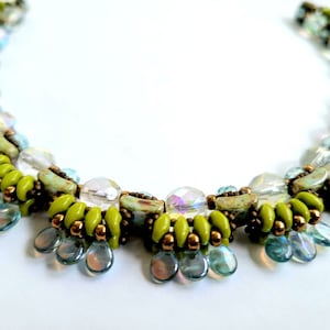 Puede incluir: Un collar de cuentas con una paleta de colores vibrantes. Presenta una combinación de cuentas verde lima, transparentes y azul iridiscente. El diseño incluye colgantes en forma de lágrima y una estructura detallada y en capas.