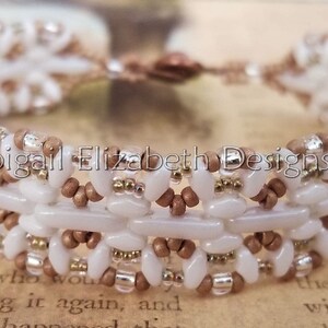 May include: A handmade bracelet featuring a woven design with white, brown, and gold beads. The bracelet has a secure clasp and the text "Abigail Elizabeth Designs" is visible. This piece of jewelry is suitable for everyday wear.