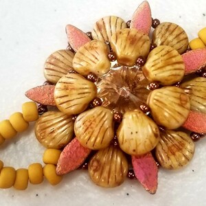 May include: A close-up of a beaded bracelet featuring a flower design. The flower is made of tan shell-shaped beads with brown stripes, pink leaf-shaped beads, and a central crystal. The bracelet has yellow beads.
