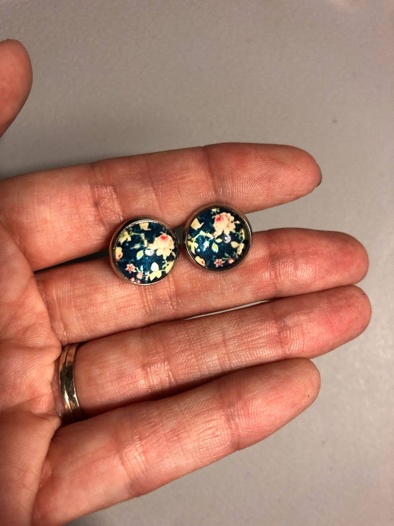 May include: Two round stud earrings with a floral design. The earrings feature a dark teal background with a pattern of small white and pink flowers. The earrings are set in silver-colored metal.