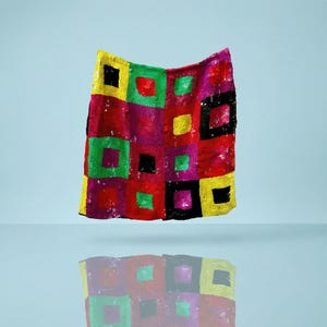 May include: A colorful, sequined textile with a patchwork design. The squares are in shades of red, yellow, green, purple, and black. The textile is displayed against a light blue background, with its reflection visible below.