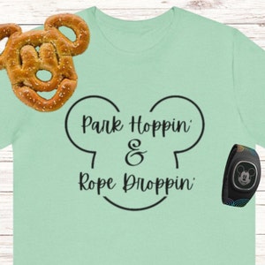 Könnte beinhalten: Ein hellgrünes T-Shirt mit einem schwarzen Mickey-Maus-Kopf-Design. Der Text "Park Hoppin' & Rope Droppin'" befindet sich innerhalb des Mickey-Maus-Kopfes. Ein schwarzes MagicBand mit einem Regenbogenmuster und einem Mickey-Maus-Kopf befindet sich in der rechten unteren Ecke des Bildes.