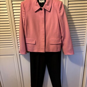 May include: A two-piece suit consisting of a light pink long-sleeve jacket with a collar, a single front button, and two flap pockets, paired with solid black trousers. The jacket features a label inside the collar with the text KASPER.