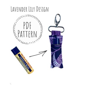 Könnte beinhalten: Ein lippenbalsam-Halter aus Stoff mit violettem und weißem Muster und einem silbernen Schlüsselanhänger. Der Halter ist für einen Lippenbalsam-Stift in Standardgröße konzipiert. Der Text "PDF PATTERN" ist in einem Kreis auf weißem Hintergrund geschrieben.