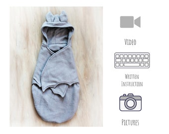 May include: A gray bat swaddle with a hood and bat wings. The swaddle is on a wooden surface. The image also includes icons for video, written instructions, and pictures.