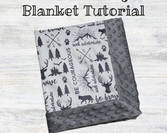 May include: A white and gray blanket with a self-binding edge. The blanket has a pattern of black and white illustrations of trees, mountains, and compass symbols. The text "Be Courageous", "Seek Adventure", and "Be Brave" is printed on the blanket.