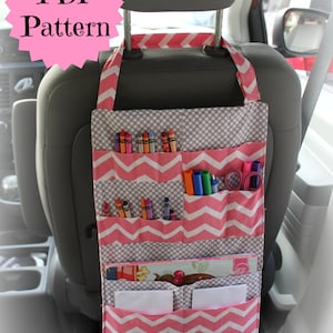 May include: Pink and gray fabric car seat organizer with multiple pockets for storing items like crayons, markers, and paper. The organizer hangs from the headrest of a car seat.