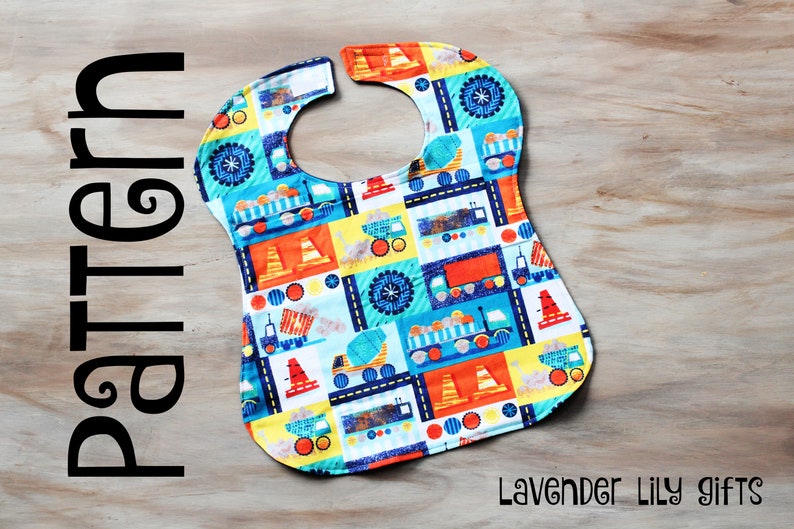 May include: A white bib with a colorful pattern of construction trucks, tires, and cones. The bib has a neck opening and is made of fabric. The text "Lavender Lily gifts" is visible in the bottom right corner of the image.