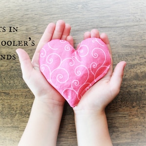 May include: A pink heart-shaped fabric pouch with a white swirl pattern, held in the hands of a child. The text "Fits in Preschooler's Hands" is visible above the pouch.