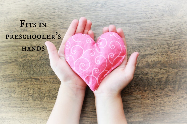 May include: A pink heart-shaped fabric pouch with a white swirl pattern, held in the hands of a child. The text "Fits in Preschooler's Hands" is visible above the pouch.