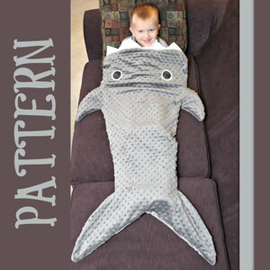 May include: A gray shark-shaped blanket with a textured surface and white accents. The blanket has fins and a tail, and is designed to be worn. The word "PATTERN" is printed vertically on the left side.