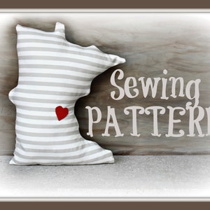 May include: A sewing pattern for a pillow shaped like the state of Minnesota. The pillow is gray and white striped with a red heart in the center. The words "Sewing Pattern" are written in white on a wood background.
