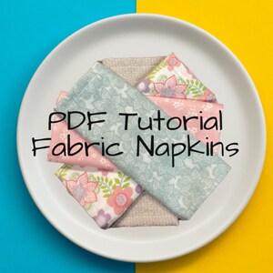 May include: A white plate with four fabric napkins folded in a decorative pattern. The napkins are in shades of pink, blue, and white with floral patterns. The text "PDF Tutorial Fabric Napkins" is written in black on the plate.