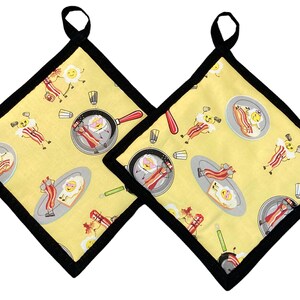 Op de afbeelding: Twee gele ovenwanten met een zwarte rand. De ovenwanten hebben een herhalend patroon van cartoon-eieren en spek.