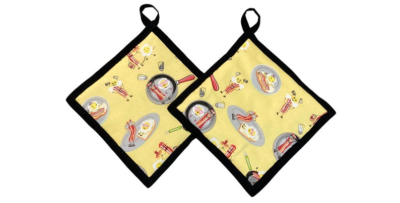 Op de afbeelding: Twee gele ovenwanten met een zwarte rand. De ovenwanten hebben een herhalend patroon van cartoon-eieren en spek.