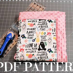 May include: A PDF pattern for a baby blanket with a pink minky backing and a white cotton front featuring a colorful print with animals and the text "I love you to the moon and back you make my world a better place".