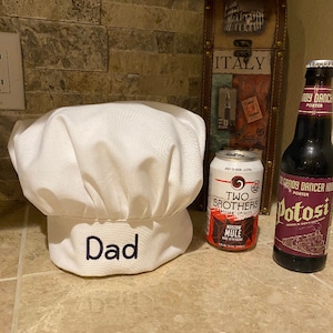Puede incluir: Un gorro de chef blanco con la palabra "Papá" bordada en el borde. Dos latas de cerveza, una con la etiqueta "Two Brothers Artisan Spirits Moscow Mule" y la otra con la etiqueta "Candy Dancer Porter Potosi", están en una encimera junto al gorro.