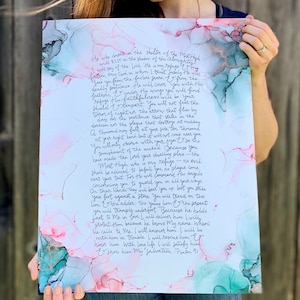 Puede incluir: Una pintura de acuarela con un fondo azul verdoso y rosa. La pintura presenta un extracto manuscrito del Salmo 91, un pasaje de la Biblia sobre encontrar refugio en Dios. El texto est&aacute; escrito en una escritura cursiva fluida.