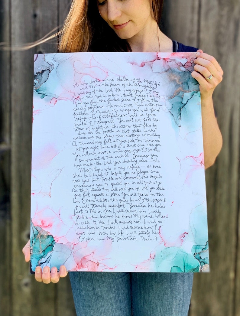 Puede incluir: Una pintura de acuarela con un fondo azul verdoso y rosa. La pintura presenta un extracto manuscrito del Salmo 91, un pasaje de la Biblia sobre encontrar refugio en Dios. El texto est&aacute; escrito en una escritura cursiva fluida.