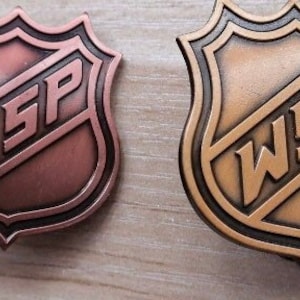 May include: Two bronze-colored hockey-style pins with a black outline. The pins feature the letters "WSP" in a slanted font.