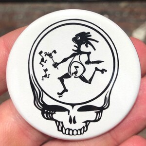 May include: A white button with a black and white image of a skull with a dancing figure inside. The skull has a wavy design on the top and a row of teeth on the bottom.
