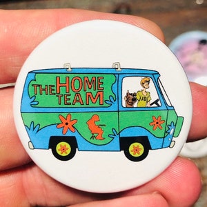 May include: A round button with a white background featuring a cartoon image of the Mystery Machine van from Scooby-Doo. The van is green, blue, and yellow with the words "The Home Team" written on the side. A cartoon character is driving the van.