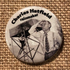 May include: A white button with a black and white photo of a man standing on a platform, looking up at the sky. The text "Charles Hatfield rainmaker" is printed in black around the edge of the button.