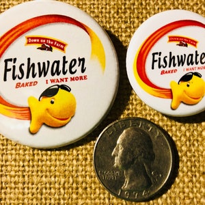 May include: Two Fishwater brand baked snack crackers buttons. The larger button features a yellow fish with sunglasses and the text "Fishwater Baked I Want More". The smaller button features a yellow fish and the text "Fishwater Baked I Want More".