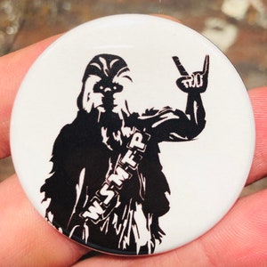 May include: Black and white button featuring a silhouette of a character with a furry head and a peace sign, wearing a sash with the text "WSMFP".