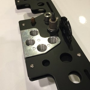 May include: A black metal reloading block with multiple holes and a silver metal component with five holes. The block has a small black cylinder and a silver metal component with a threaded top.