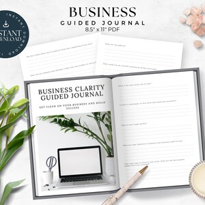 May include: An open business clarity guided journal with a white background and black text. The journal is open to a page with the title "Business Clarity Guided Journal" and the text "Get clear on your business and build success". The journal is on a white surface with a green plant, a laptop, a white mug, a whisk, and a pen.