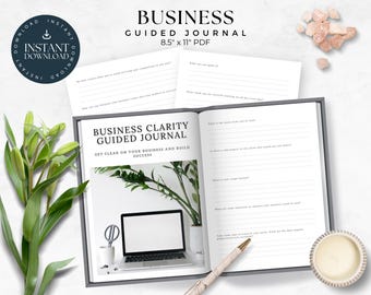 May include: An open business clarity guided journal with a white background and black text. The journal is open to a page with the title "Business Clarity Guided Journal" and the text "Get clear on your business and build success". The journal is on a white surface with a green plant, a laptop, a white mug, a whisk, and a pen.