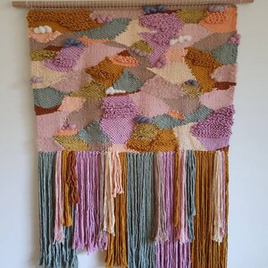 May include: A colorful woven wall hanging with a geometric pattern in shades of pink, orange, yellow, and green. The hanging has a fringe border and is mounted on a wooden dowel.