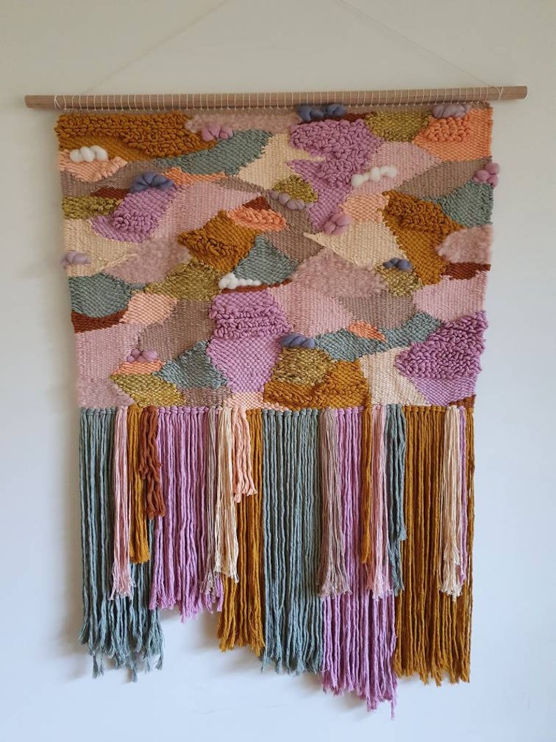 May include: A colorful woven wall hanging with a geometric pattern in shades of pink, orange, yellow, and green. The hanging has a fringe border and is mounted on a wooden dowel.