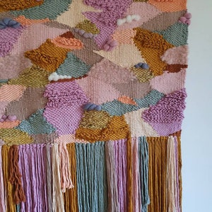 May include: A colorful woven wall hanging with a geometric pattern and a fringe border. The hanging features shades of pink, orange, yellow, green, and brown.