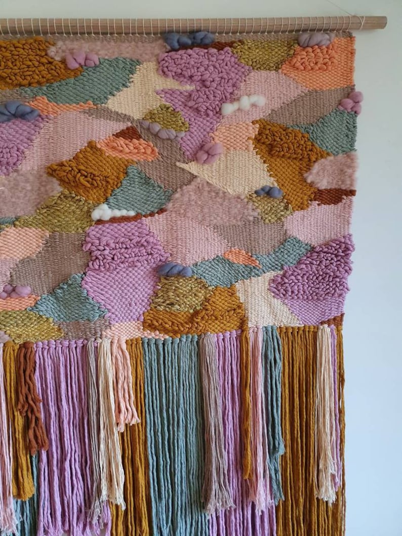 May include: A colorful woven wall hanging with a geometric pattern and a fringe border. The hanging features shades of pink, orange, yellow, green, and brown.